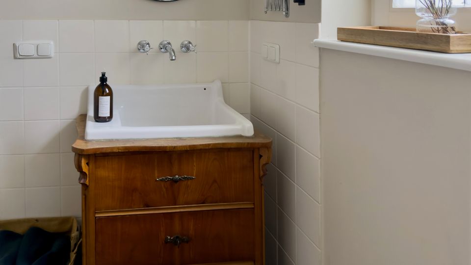 Helles Badezimmer mit wandhängendem Waschbecken auf einem antiken Holzschrank, runden Wandspiegeln und weißen Fliesen bis zur halben Wandhöhe. Rechts ein großes Fenster mit Fensterbank und Tablett mit Vase, darunter grauer Fliesenboden.