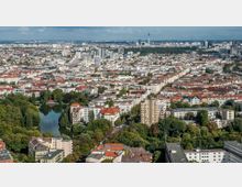 Luftaufnahme einer Stadt mit dichter Bebauung und zahlreichen roten Dächern, umgeben von grünen Parks und einem kleinen See. Im Hintergrund ist der Berliner Fernsehturm sichtbar, der sich über die Skyline erstreckt.