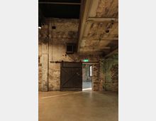 Industriehalle mit grober Backsteinwand, einer schwarzen Holztür im Scheunentor-Stil und freiliegenden Rohren an der Decke. Durch die geöffnete Tür sichtbar ist ein angrenzender Raum mit großen Fenstern.