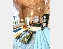 Ein gemütlicher Loungebereich in einem Raum mit Backsteinwänden und einer Holzdecke. Der Raum ist eingerichtet mit einem blaugrauen Sofa, zwei ockerfarbenen Sesseln, einem Couchtisch aus Glas und Metall, sowie Regalen und Schränken im Hintergrund; ein großes Fenster mit schwarzem Rahmen lässt Tageslicht herein.