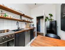 Helle, schmale Küche mit weißer Fliesenrückwand, Holzarbeitsplatte, schwarzer Spüle und offenem Wandregal mit Gläsern und Geschirr. Rechts steht ein schwarzer Kühlschrank neben einer Tür zum Flur; im Vordergrund ist ein Holztisch zu sehen.