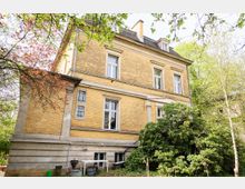 Gelbes Backsteingebäude mit weißen Fensterrahmen, umgeben von Bäumen und Sträuchern in einem grünen Garten. Das Gebäude hat zwei Etagen sowie ein Dachgeschoss mit kleinen Fenstern und ist von Frühlingslaub umgeben.