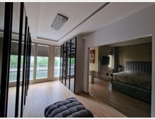 Ein moderner Ankleideraum mit Holzboden, bodentiefen Fenstern mit Blick auf einen Fluss und hellen Wänden. Auf der rechten Seite befindet sich eine große Spiegelwand, die ein Schlafzimmer mit einem grauen gepolsterten Bett reflektiert.