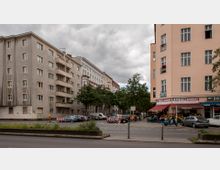 Straßenecke in einer urbanen Umgebung mit mehrstöckigen Wohngebäuden; links ein grauer Bau mit Balkonen, rechts ein beiges Gebäude mit Geschäften im Erdgeschoss, darunter ein Lebensmittelgeschäft und ein Imbiss. Im Vordergrund parkende Autos, Fahrräder und ein kleiner begründerter Bereich; Bäume säumen die Straße.