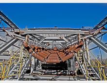Industrielle Förderanlage mit rostigen Metallrollen, eingebettet in eine Struktur aus Stahlträgern und Geländern, unter freiem Himmel bei blauem Himmel. Im Hintergrund sind weitere Industrieelemente erkennbar.