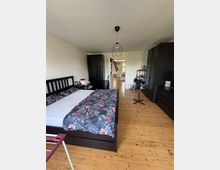 Schlafzimmer mit Holzdielenboden, großem Doppelbett mit dunklem Holzrahmen und gemusterter Bettwäsche. An den Wänden stehen mehrere schwarze Kleiderschränke; in der Mitte hängt eine geometrische Deckenlampe, und eine offene Tür führt in einen hellen Flur.