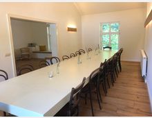 Heller Raum mit einem langen weißen Tisch, umgeben von dunklen Holzstühlen, auf einem Holzboden. Im Hintergrund befindet sich ein Durchgang zu einem weiteren Bereich mit Korbsessel und Sitzbank; ein Fenster mit Blick auf grüne Vegetation lässt natürliches Licht herein.