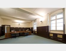 Innenraum eines Gerichtssaals mit einer hölzernen Richterbank, drei Stühlen dahinter und weiteren Stühlen davor. Die Wände sind teilweise mit dunkler Holzvertäfelung und großen Fenstern versehen, die Tageslicht hereinlassen.