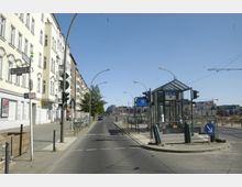 Straßenszene in einer Stadt mit mehrstöckigen Wohnhäusern auf der linken Seite und einem Eingang zur U-Bahn-Station „Schwartzkopffstraße“ der Linie U6 auf der rechten Seite. Der Eingang ist von einer Glaskonstruktion überdacht, daneben befinden sich Straßenschilder und Ampeln. Im Hintergrund sind Bauflächen und moderne Gebäude zu sehen.