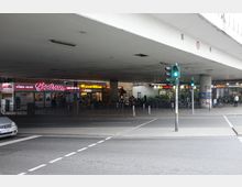 Straßenszene unter einem Betonbrückenbauwerk mit Geschäften, darunter ein Döner-Imbiss ("Bodrum") und ein Asia-Restaurant ("Asia24"). Im Vordergrund sind eine Ampel, ein Zebrastreifen und geparkte Fahrräder zu sehen.