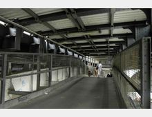 Ein überdachter Fußgängersteg mit Metallgittern an den Seiten, einer Betonrampe und einer Stahlkonstruktion mit sichtbaren Trägern an der Decke. Im Hintergrund sind Personen zu sehen, die den Steg nutzen.