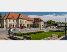 Historisches Bahnhofsgebäude mit heller Fassade und roten Ziegeldächern, umgeben von einem modernen Platz mit Bänken, Grünflächen und kleinen Wegbegrenzungen. Im Hintergrund sind eine Fahrradstation, mehrere Autos und Bäume zu sehen.