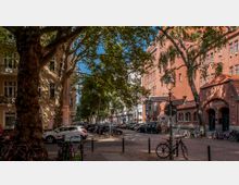 Straßenszene mit Kopfsteinpflaster, flankiert von historischen Gebäuden aus rotem Backstein und hell verputzten Wohnhäusern mit großen Fenstern. Im Vordergrund stehen Bäume, Fahrräder und parkende Autos, während Straßenschilder „Roscherstraße“ und „Sybelstraße“ anzeigen.