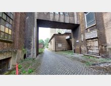 Industriegelände mit alten Gebäuden aus Backstein und Beton. Kopfsteinpflaster führt durch eine überdachte Passage, flankiert von rostigen Türen und vergilbten Fenstern, mit Blick auf eine grüne Umgebung im Hintergrund.