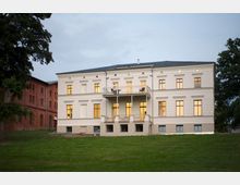 Historisches, zweistöckiges Gebäude mit heller Fassade und regelmäßigen Fenstern, das von einem gepflegten Rasen umgeben ist. Im Hintergrund befindet sich ein angrenzendes rotes Backsteingebäude; ein Baumrahmen flankiert die Szenerie.