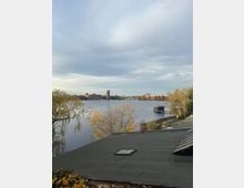 Blick über einen breiten Fluss oder See mit bewölktem Himmel; am gegenüberliegenden Ufer stehen mehrere Backstein- und moderne Gebäude sowie eine Brücke. Im Vordergrund sind ein flaches Dach, herbstliche Bäume und rechts ein Steg mit einem kleinen Bootshaus am Ufer zu sehen.