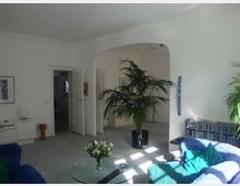 Wohnzimmer mit hellen Wänden, breitem Torbogen und mehreren Zimmerpflanzen in schwarzen Töpfen. Die Einrichtung umfasst ein blaues Sofa, einen Glastisch mit Blumenstrauß, ein weißes Sideboard und ein Regal mit CDs.