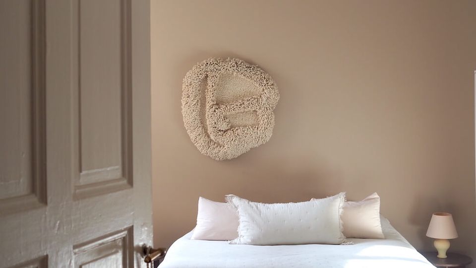 Minimalist bedroom with beige walls and a white bed, featuring a textured wall sculpture above the headboard and a large round paper pendant light on the ceiling. A low upholstered bench at the foot of the bed holds books and decor, with a small bedside table and lamp beside an open paneled door.