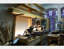 Werkstatt mit Holzbearbeitungsausstattung, einschließlich einer Gehrungssäge auf einer Werkbank, Regalen mit Holzplatten und Werkzeugen sowie Fenstern im Hintergrund, die einen leicht industriellen Charakter des Raums andeuten.