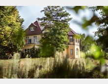 Zweistöckiges gelbes Gebäude mit rotem Dach und Fachwerkelementen, umgeben von Bäumen und einem leicht verwilderten Garten. Im Vordergrund befindet sich eine Wiese mit hohen Gräsern.