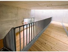 Ein minimalistischer Innenraum mit Betonwänden und einer Decke aus Sichtbeton, einer Holzplattform mit Metallgeländer, sowie einer bodentiefen Glasfront, die Tageslicht hereinlässt.