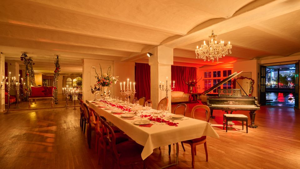 Großer, warm beleuchteter Veranstaltungsraum mit Holzboden, niedriger Decke und roten Vorhängen, in der Mitte ein lang gedeckter Esstisch mit Kerzenleuchtern. Rechts steht ein schwarzer Flügel unter einem Kronleuchter, dahinter große Fenster und eine offene Tür mit Blick auf die nächtliche Straße.