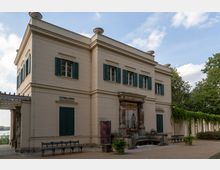 Zweistöckiges klassizistisches Gebäude mit cremefarbener Fassade und grünen Fensterläden, flankiert von einer überdachten Kolonnade mit Pflanzenbewuchs. Im Vordergrund eine kleine Brunnenanlage mit Statuen sowie Bänke auf einem Kiesweg.