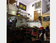 Ein kleines, dicht eingerichtetes Werkstattzimmer mit einem Arbeitstisch, Regalen voller Werkzeuge und Materialien sowie Wanddekorationen, darunter Bilder und Tiermotive. Eine Spüle, zahlreiche Lampen und Farben auf rollbaren Wagen und Regalen verleihen dem Raum eine funktionale Atmosphäre.