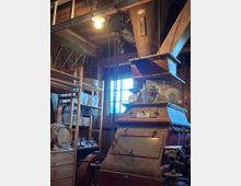 Innenraum einer rustikalen Werkstatt oder Mühle mit Holzbalken, Fenster und hängendem Kettenzug unter der Decke. Im Vordergrund steht eine große, alte Metallmaschine, umgeben von Regalen mit Werkzeugen und Gegenständen sowie einem Holzfass.