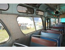 Innenraum eines alten Busses mit abgenutzten Sitzbänken in brauner und blauer Farbe. Durch die großen Fenster sind mehrere geparkte Fahrzeuge und eine grüne Wiese zu sehen.