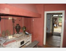 Ein historischer Innenraum mit rötlichen Wänden, einem gemauerten Herd aus Ziegelsteinen und traditionellen Gegenständen wie einem Samowar und Flechtwerk-Schuhen auf der Herdfläche. Im Hintergrund öffnet sich eine Tür zu einem gepflasterten Außenbereich mit Blick auf eine Holzzaun-Umgebung.