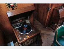 Antiker Plattenspieler in einem hölzernen Gehäuse mit geöffneter Abdeckung, umgeben von Holzmöbeln und Parkettboden; im Hintergrund eine grüne Stuhllehne und eine Tür.