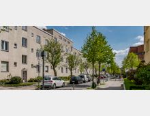 Wohnstraße mit mehrstöckigen, hellen Wohngebäuden auf der linken Seite und einer Baumreihe, die entlang der Straße verläuft. Rechts daneben befindet sich ein Fußweg mit Büschen, parkende Autos säumen die Fahrbahn.