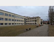 Großer Innenhof vor einem mehrstöckigen, hellbeigen Schul- oder Verwaltungsgebäude mit vielen rechteckigen Fenstern und einem verglasten Treppenhaus. Daneben verläuft ein gepflasterter Weg mit Bänken und einzelnen Bäumen auf einer Rasenfläche unter bewölktem Himmel.