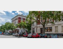 Straßenszene mit Altbauten im gründerzeitlichen Stil, erkennbar an roten Backsteinfassaden und weißen Stuckverzierungen. Im Vordergrund parken Autos entlang der Straße, während Bäume teils den Gehweg und die Fassaden beschatten; ein Radfahrer fährt auf der Straße.