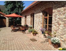 Ziegelhof mit einer Terrasse, auf der ein Holztisch und Stühle unter einem roten Sonnenschirm stehen; daneben Blumen in Töpfen auf Backsteinstufen vor einer Hauswand aus hellem, gemischtem Backstein mit großen Fenstern. Im Hintergrund steht eine Tanne, und links ist ein Nebengebäude zu sehen.
