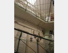 Innenansicht eines Gebäudes mit industriellem Charakter, erkennbar an Metallgeländern, Stromkästen und freiliegenden Kabeln. Eine Tür und eine Glaswand mit Tür befinden sich im unteren Bereich, während ein Oberlicht Tageslicht hineinlässt.