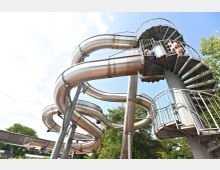 Metallische Wasserrutsche mit mehreren Windungen und Kurven, die von einem spiralförmigen Treppenturm aus zugänglich ist. Der Hintergrund zeigt blauen Himmel und grüne Bäume.