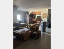 Wohnzimmer in einem offenen Grundriss mit einer großen braunen Eckcouch, einem Beistelltisch mit Dekoration, und einem Kaminofen in der Ecke. Im Hintergrund ein Essbereich mit Esstisch und Stühlen sowie dekorativer Wandkunst.