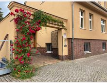 Gelbes Wohnhaus mit braunem Klinkersockel, einem überdachten Eingangsbereich und außen wachsenden roten Rosen, die entlang einer Rankhilfe bis zum Eingang hochklettern. Der Boden vor dem Haus ist mit Kopfsteinpflaster ausgelegt.