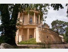 Klassizistisches Rundgebäude aus gelbem Stein mit Säulen, Rundbögen und Fenstern; im Vordergrund Bäume und Büsche, im Hintergrund bewaldete Umgebung.