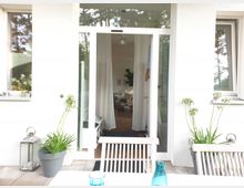 Außenansicht eines modernen Hauses mit weißer Fassade und großen Glasfenstern. Im Vordergrund steht ein Holztisch auf einer Terrasse, flankiert von Blumentöpfen und einer Laterne, während eine geöffnete Tür Einblick in ein gemütlich eingerichtetes Wohnzimmer gewährt.