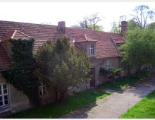 Langgestrecktes Gebäude mit roten Ziegeldächern und verputzten Fassaden, von Bäumen und Efeu teilweise umgeben. Ein gepflasterter Weg verläuft entlang des Hauses, das von einer grünen, parkähnlichen Umgebung umgeben ist.