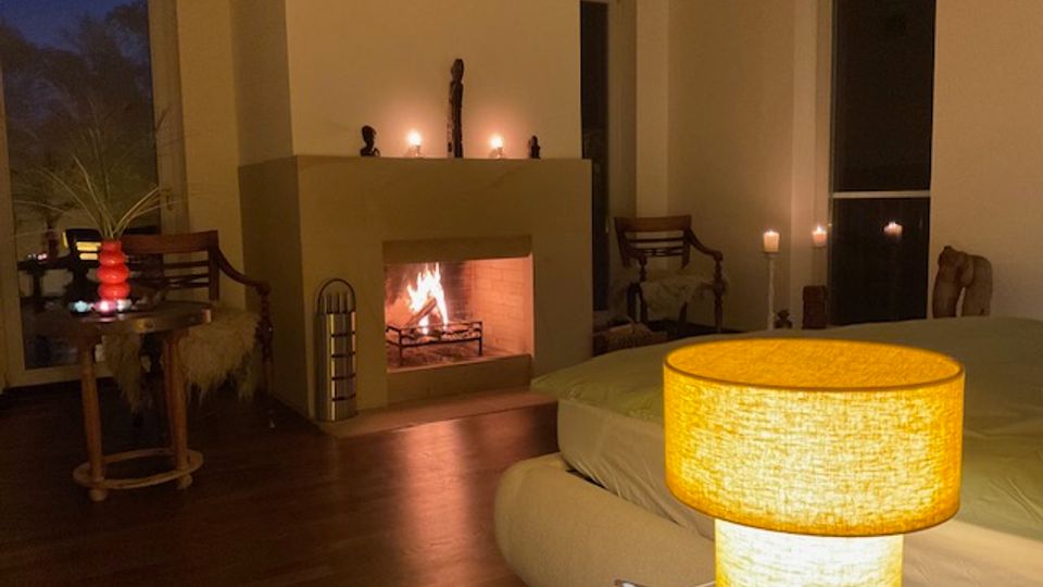 Gemütliches Wohnzimmer mit Holzboden, zentralem Kaminfeuer und Kaminsims mit Kerzen und kleinen Skulpturen. Große Fensterfronten, ein niedriges Sofa sowie ein Beistelltisch und Stühle ergänzen die warm beleuchtete Einrichtung.