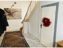 Eingangsbereich eines Hauses mit weiß gefliesten Böden, einer Holztür, die mit einem roten Beerenkranz dekoriert ist, und einer Treppe mit grauem Geländer. An der Wand hängt ein Kunstwerk, der Boden ist mit einem gemusterten Läufer bedeckt.