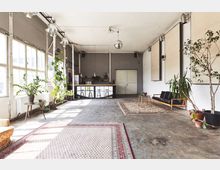 Großer, heller Raum mit Betonboden und großen Fenstern auf der linken Seite, der mit Topfpflanzen, Teppichen und einer schwarzen Sitzgruppe möbliert ist. Im Hintergrund befindet sich eine kleine Bar mit hohen Hockern und einer Discokugel an der Decke.