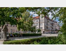 Ein Platz mit Kopfsteinpflaster und grüner Bepflanzung, mit einer Steinfigur im Vordergrund. Im Hintergrund sind mehrere historische Wohn- und Geschäftshäuser mit heller Fassade und roten Dächern zu sehen, umgeben von Bäumen und Menschen, die auf Bänken sitzen.