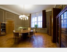 Ein elegantes Besprechungszimmer mit einem runden Holztisch und sechs Stühlen in der Mitte, hohen Fenstern mit weißen Vorhängen, braunen Verdunkelungsvorhängen und einem Kronleuchter. Holzmöbel, darunter Bücherregale und eine geschlossene Tür im klassischen Stil, ergänzen den Raum, der einen Parkettboden hat.