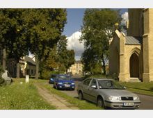 Straßenszene in einer Kleinstadt mit parkenden Autos auf einem Seitenstreifen, flankiert von Bäumen und Rasenflächen. Auf der rechten Seite ist ein Teil einer Backsteinkirche mit einem Spitzbogenportal sichtbar, im Hintergrund Wohnhäuser und ein größeres gelbes Gebäude.