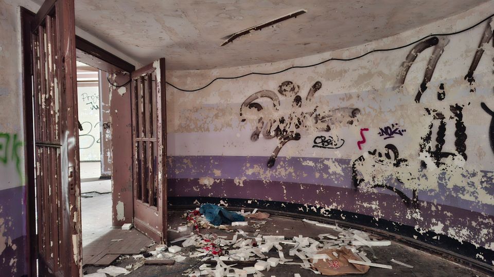 Innenraum eines verlassenen, heruntergekommenen Gebäudes mit abblätternder Farbe, Graffiti an den Wänden und violetten Wandstreifen. Offene, beschädigte Türen führen in einen weiteren Raum; der Boden ist mit Schutt, Papier und Trümmern bedeckt.
